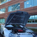 A silver sports car with an open hood parked in front of a modern office building, showcasing its engine during a cars+coffee event.
