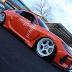 Low angle view of a bright orange sports car with custom graphics and white wheels parked at a cars+coffee event.