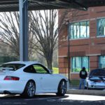 A white sports car parked under a canopy with two men standing nearby, talking at a Cars+Coffee event, in front of a brick building on a sunny day.