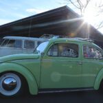 A vintage green Volkswagen Beetle parked outdoors at a cars+coffee event, with sunlight glaring from behind, showcasing its pristine condition and retro design.