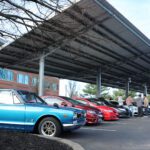 Row of vintage cars, including a blue Datsun 510, parked under a solar panel carport beside an office building for a Cars+Coffee event.