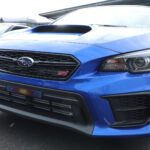 Blue Subaru WRX STI parked at a Cars+Coffee event, showing a close-up of its front fascia with distinctive grille and logo.