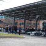 People at an outdoor "Cars+Coffee" event under a large shelter, browsing and discussing various parked cars.