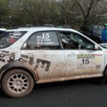 A muddy Subaru rally car with race decals parked at a cars+coffee event, with two people standing by it in a casual setting.