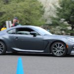 A gray sports car driving fast around a cone-marked course with blurred background indicating motion at a cars+coffee event.