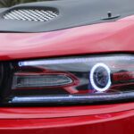 Close-up of a red car's headlight with "hellcat" logo illuminated at a cars+coffee event.