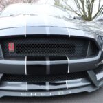 Front view of a black Shelby Mustang with white stripes parked at a cars+coffee event, displaying its grille and emblem.
