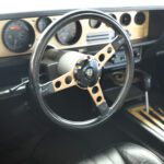 Interior view of a vintage car showing the steering wheel, dashboard, and gauges, with a focus on the steering wheel during a cars+coffee event.