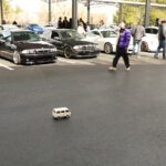 A small remote-controlled car drives on a parking lot surrounded by people and full-sized cars during a Cars+Coffee event, including several BMWs.