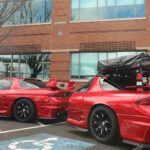 Two red sports cars parked side by side at a cars+coffee event, the one on the right equipped with a roof rack and cargo bag, in a parking lot with a brick building in the background