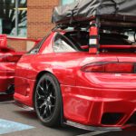 Two red sports cars parked side by side, one with roof cargo box, in a parking lot near buildings during a cars+coffee event.