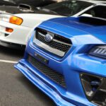 Close-up view of a blue Subaru car with its grille and hood visible, parked next to a white car at a cars+coffee event, focusing on performance vehicle features.