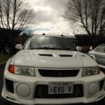 Front view of a white Mitsubishi Lancer Evolution V with custom enhancements parked outdoors at a cars+coffee event under a cloudy sky.