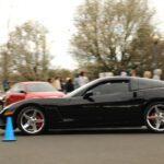A black sports car in motion at a Cars+Coffee event with a blurred background of spectators and other vehicles.