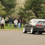 A black sports car drives past a group of spectators at a cars+coffee event on a tree-lined street.