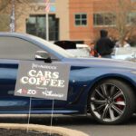 Blue car with a "cars+coffee" event sign in front at a gathering, with people and flags in the background.