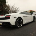 White Lamborghini Gallardo parked on a street on a cloudy day, viewed from the rear left side, near a cars+coffee event.