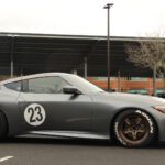 A matte gray sports car with the number 23 on the side, parked in a lot at a cars+coffee event with other cars and bare trees in the background.