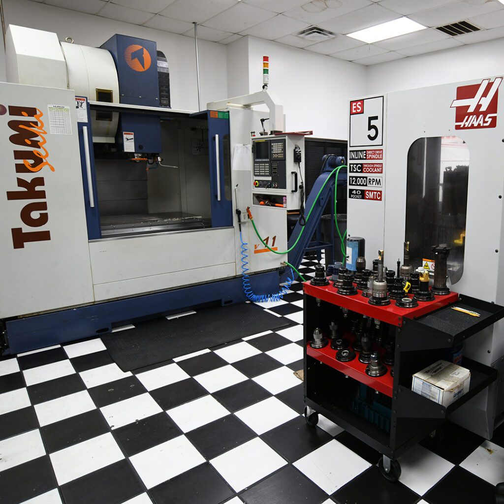 An industrial workplace featuring two different CNC machines with a checkered floor and a tool cart nearby, often used for practical lectures.